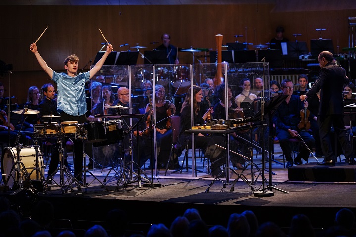 To the left of the image, Jordan is wearing an electric blue silk shirt, holding drumsticks up above his head. In front of him is a multi-percussion drum setup, including bongos, tomtoms and cymbals. Central, is another multi-percussion setup, with temple blocks, woodblocks, castinets, and a car brake drum. Behind him, clear acoustic screens separate him from the violins of the BBC Philharmonic Orchestra, with Mark Wigglesworth to the right.
