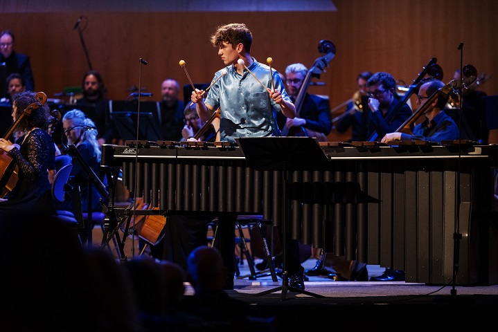 Jordan is wearing an electric blue silk shirt, standing centrally in the image, in front of a five octave marimba. He is holding two mallets in each hand, with their heads up at shoulder height. Behind him are the lower strings and brass of the BBC Philharmonic Orchestra, and he is facing towards conductor Mark Wigglesworth, who is out of frame to the left.