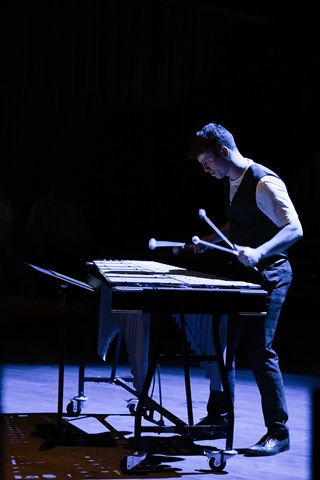 The image is in portrait orientation and shows a side-on view of Jordan at a vibraphone. He is wearing a dark blue waistcoat over a white t-shirt, and dark blue trousers with black shoes. He has two mallets in each hand - all slightly blurred with the movement. The background is dark, but the overhead lights provide a blue hue to reinforce the theme of the music he is playing: MoonVibe by Krystian Skubała.