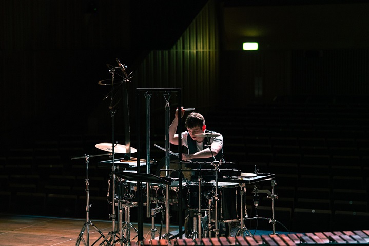 The photo shows Jordan on stage at the Royal Birmingham Conservatoire, during his BMus final year recital. He is wearing a dark blue waistcoat over a white t-shirt, with a drumstick in each hand - one above his head, the other playing in front of him. Playing Tobias Broström's 