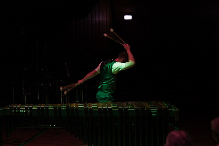 The photo shows a darkened stage, with Jordan standing behind a MarimbaOne marimba, and wearing a waistcoat over a white t-shirt. It is an action shot, with Jordan having just played a final descending glissando, so is spinning away from the camera, to his left. He has two mallets in each hand, his right hand raised above his head and his left hand thrown out behind him. These were the final notes of his BMus final year recital at the Royal Birmingham Conservatoire.