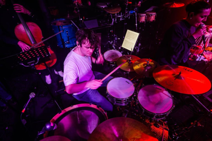 Jordan Ashman is pictured centrally, playing drumkit during a soundcheck for his 'Convergence' event. He is wearing bluejeans and a white T-shirt. To the right of the photo is Grace Conner, vocalist, wearing a dark grey sweatshirt and holding a microphone to her mouth. Behind them is a large array of instruments, including a cello, bass drum, cymbals, and bongos.