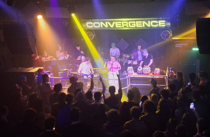 The still image shows Jordan Ashman's jungle band on stage at Mama Roux's club. The front line from left to right consists of Jordan on drumkit wearing a white T-shirt, vocalist Grace Conner wearing a pink top, and Dal the Tabla Jedi, wearing all black. Behind them is a string quartet (standing), then two synth / percussionists one of whom is wearing a frog hat. Above the stage is an electronic sign showing the word 'Convergence' in yellow - the name of the gig. In the foreground, many dancers are revelling to the music.