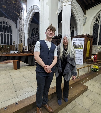 Jordan Ashman is pictured alongside and to the left of Dame Evelyn Glennie in St Neots Parish Church, after his solo recital there. Both are smiling towards the camera, standing on a small step up to a plinth that is holding a marimba. Jordan is wearing dark blue trousers and waistcoat over a white t-shirt, with brown leather shoes, and has his hands clasped in front of him. Evelyn has her long, silver grey hair worn loose, and is wearing a dark top and trousers, a light grey coat and blue suede shoes.