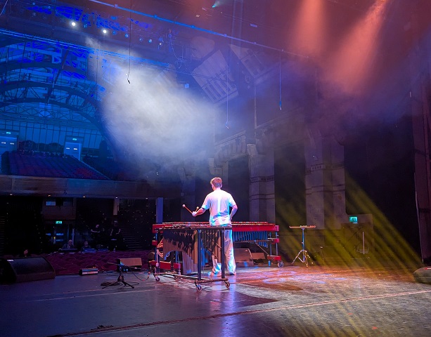 Taken from the back of the stage, Jordan is central in the image, facing away into the Cambridge Corn Exchange auditorium. He is standing in the 'v' shape formed by a five-octage marimba and a vibraphone, ready to play 'Attraction' by S&eacute;journ&eacute;. White spot lights and yellow lasers shine through the stage fog. 1400 seats in the two levels of the auditorium are empty, bar the lighting and sound team.