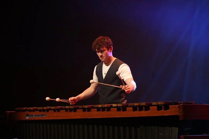 Jordan Ashman is pictured facing the camera, slightly towards the left. He is wearing a dark blue waistcoat over a plain white t-shirt. With two mallets in each hand, he is playing a Bergerault five-octave marimba, which fills the bottom third of the image. The background is blue black, with stage lights creating a blue glow to the top right.