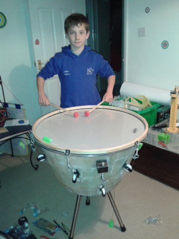 Jordan Ashman, aged 10, is standing behind his home-made timpani with his sticks resting on the skin. The timpani body is made from uncoated and opaque fibreglass, with three metal legs beneath it. The counter hoop stretching the head into tune is made from glulam (glued laminated timber), and is tightened using metal bolts attached to pullies, three of which are visible. Jordan is wearing a skyblue hoodie with the National Children's Orchestra logo and his name embroidered on the chest. The room has off-white walls and beige carpet, with the internal door wide open. There is lots of clutter surrounding him, including wooden offcuts, a tape measure, pencil, and needle files.
