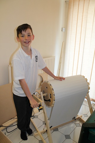 The photo shows Jordan Ashman standing alongside his home-made aeoliphone - also known as a wind machine. This instrument consists of a cylindrical slatted wooden drum, mounted horizontally on a wooden frame, with a nylon sheet pulled tightly across it. Jordan is holding a crank handle, which would rotate the cylinder against the material to create the noise of the wind. Jordan is wearing a white polo shirt with the Milton Primary School logo on its chest in blue, and with black trousers. The floor is white and grey tiles, but the background is distinctly beige - wall, radiator and vertical blinds. There is offcut wood behind him, along with a selection of woodworking tools.