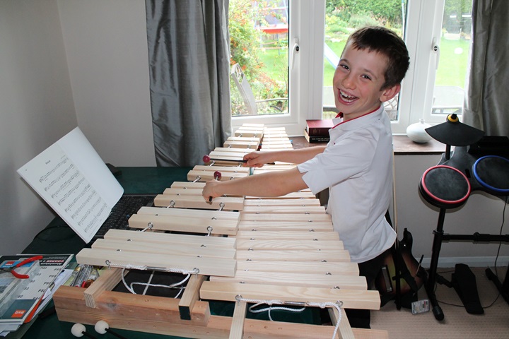 Central to the photo is a home-made xylophone, with its longest (lowest) notes closest to the camera and shorted (highest) ones further away. It is made from ash wood, and evidence of sandpaper and tools are visible on the table to the left of the photo. Jordan Ashman, aged 9, is standing to the right of the xylophone, playing it with two rubber-headed mallets, but facing the camera with a happy smile. He is wearing a white short-sleaved football shirt with red flashing around the collar, and dark trousers. A music book is propped up to the left, showing Prelude by Kevin Hathaway. To the right of the image are a Nintendo Wii guitar and drum kit. In the background, between silver curtains, a grassy garden contains a child's climbing frame, and leads to a conifer hedge in the distance.