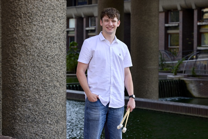 Jordan stands in the centre of the image, facing the camera. He is wearing a casual white button-down shirt and blue jeans, and looks relaxed, with his right hand in his jeans pocket, and left hand holding four mallets. The photograph is taken outside London's Barbican Centre, in front of its 'brutalist' concrete columns and interlocking ponds.