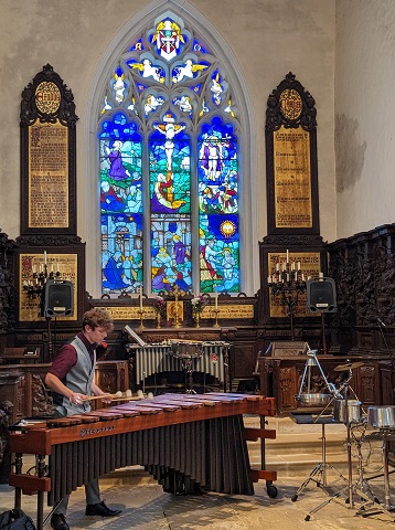 The image shows Jordan Ashman in St John's Church, Cockayne Hatley, near Sandy. He is in the lower left corner of the photo, wearing a deep red short-sleeved shirt under a silvery grey waistcoat and trousers. He is facing half towards the camera, while playing his Bergerault five-octave marimba. Behind him is his vibraphone, in front of the altar, and a tall stained glass window, through which the sun is shining. To the right of the image is an assortment of 'instruments' fashioned from pans, metal baking trays and wine cooler, all ready for his next piece.