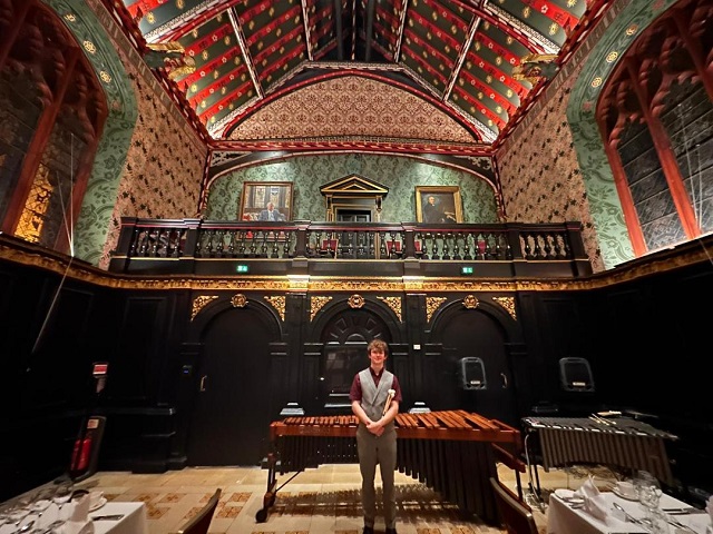 Jordan Ashman is pictured in the Queen's College Old Hall in Cambridge. He is standing centrally, in front of a marimba and vibraphone, but is only one third the height of the image, the remainder showing the gradeur of the Old Hall. The lower half of the hall has black walls with intricate gilding. Higher up is a black balcony railing, in front of rich patterned green walls, then a bright red and green striped vaulted ceiling. Triple arched windows are left and right. The bottom corners of the image show dinner tables, laid with white cloths, ready for service.