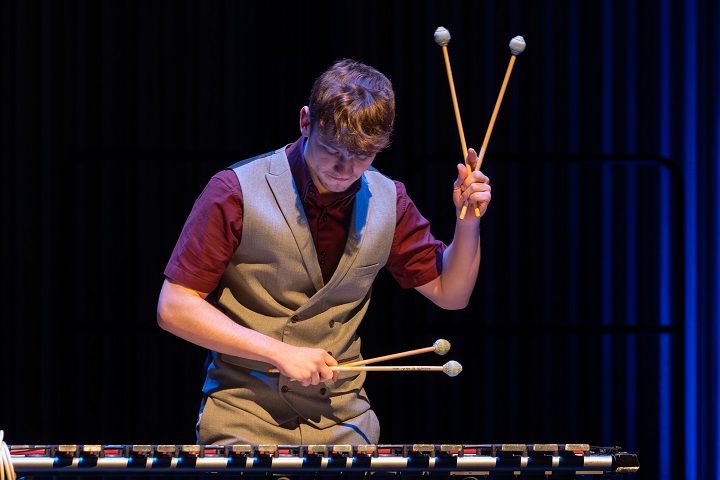 The image shows Jordan Ashman facing the camera, but with his head slightly lowered. He is wearing a siver-grey waistcoat and trousers, over a claret short-sleaved shirt with an open neck. He is playing a vibraphone, the keys of which frame the bottom of the photo. His left hand holds two mallets, raised to head height in a 'V' shape. His left hand, also holding two mallets, is held across his waist. Behind him to the right, electric blue light is reflecting off the hall's vertical acoustic baffles, fading to black towards the back of the stage.