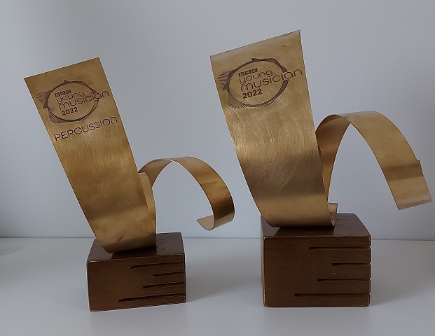 Two very similar trophies, resembling a bronze left-to-right spirals, diminishing towards their right, each mounted on a dark wooden block. Both trophies are engraved with the BBC Young Musician 2022 logo, with the smaller left one having an additional 'percussion' title.