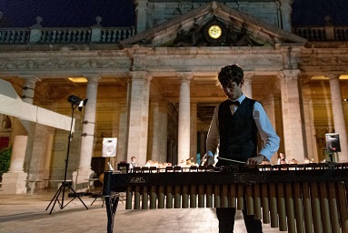 Jordan in a black waistcoat over a white shirt, playing a xylophone solo in front of the Youth Brass 2000 band, who are just visible above the xylophone. The stunning Montecatini Terme Tettuccio building towers above them all. The evening concert is outdoors, with lighting visible in shot.
